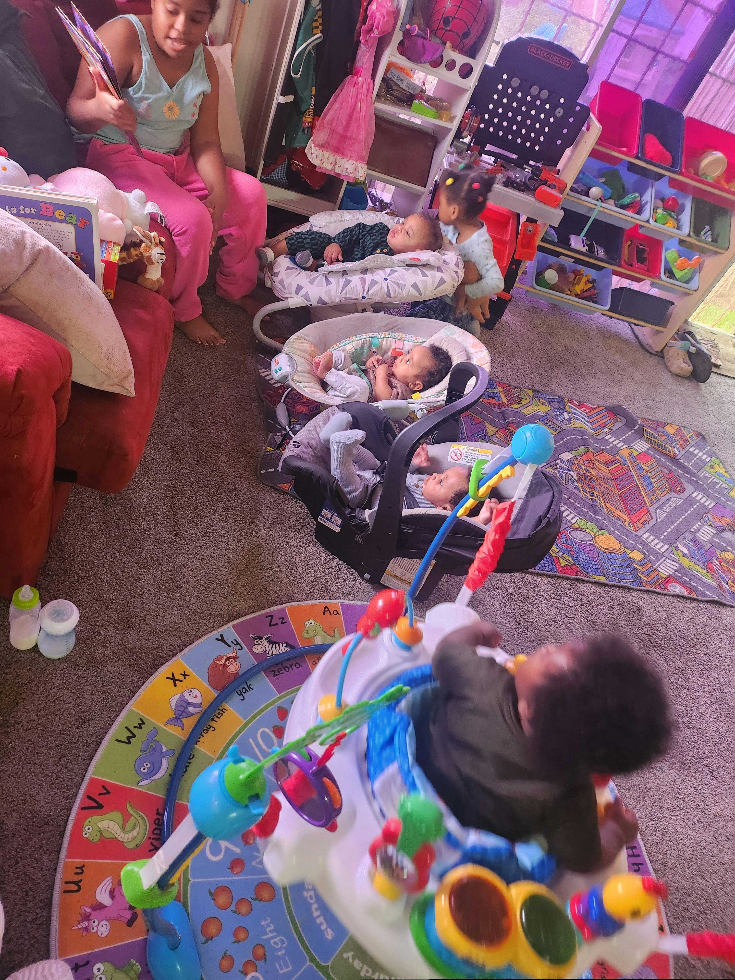 Children and babies in a room, one child reading, babies in carriers and a bouncer