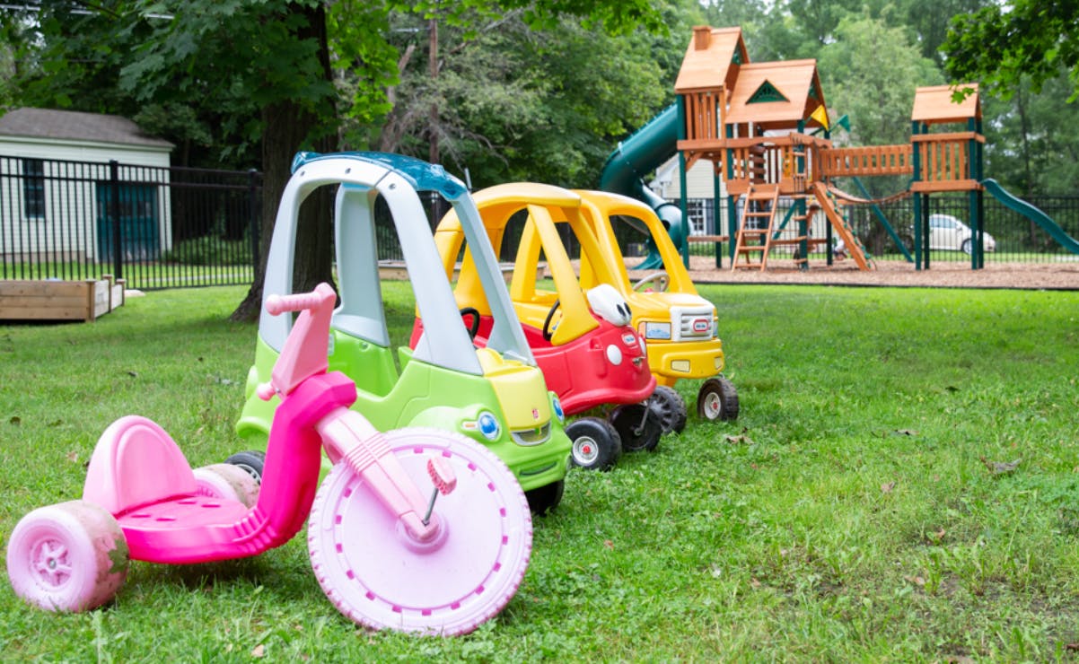 Children's tricycle and toy cars on grass with a playground in the background
