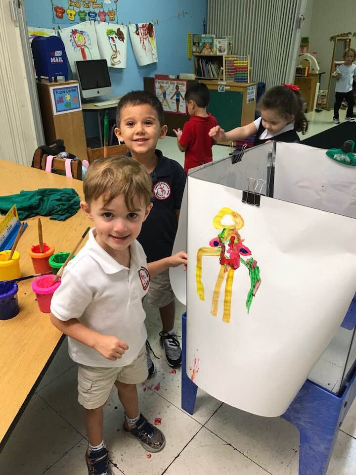 Two children smiling at the camera in a classroom with a painting