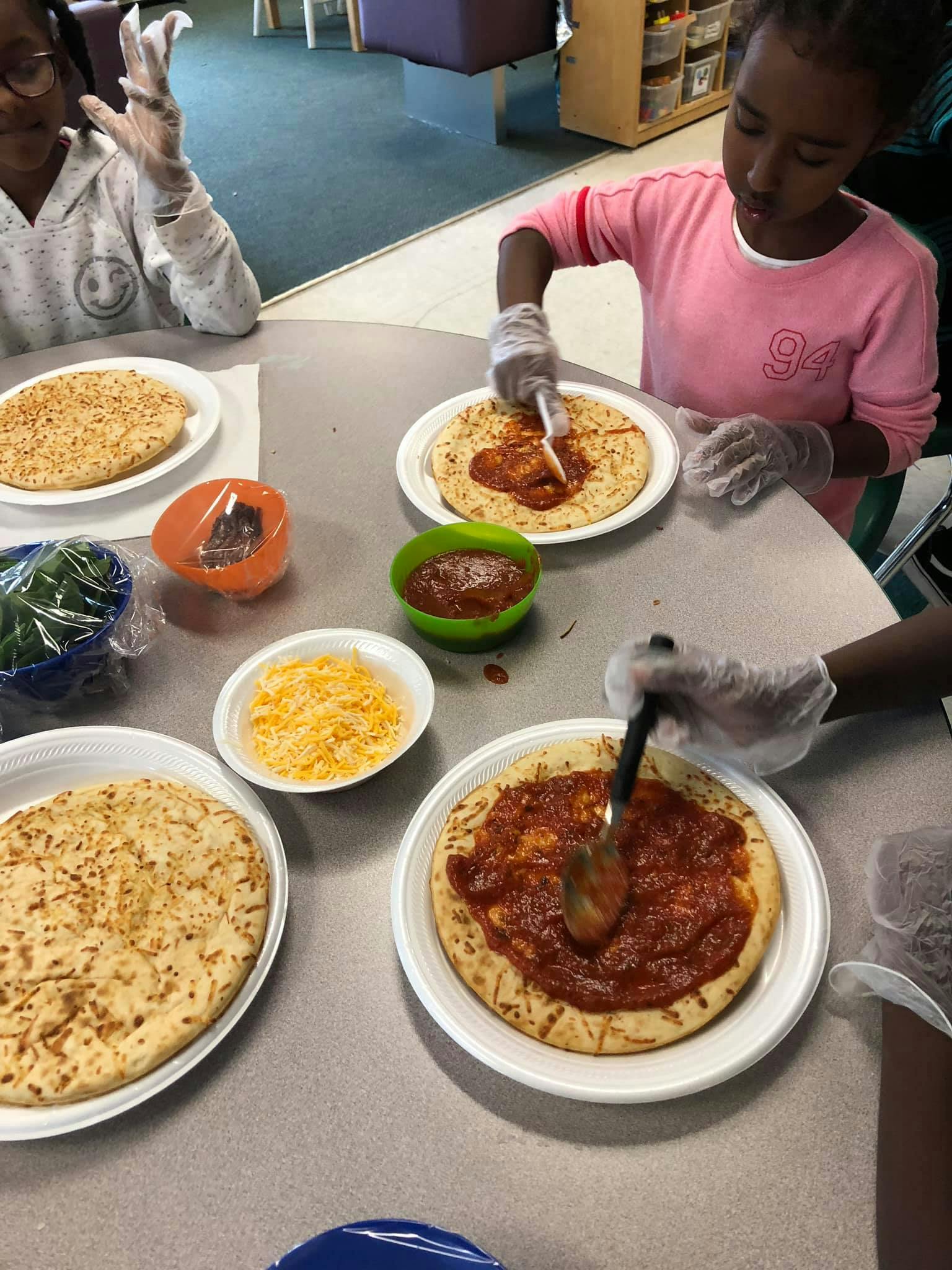 Children making pizzas, spreading sauce on crusts