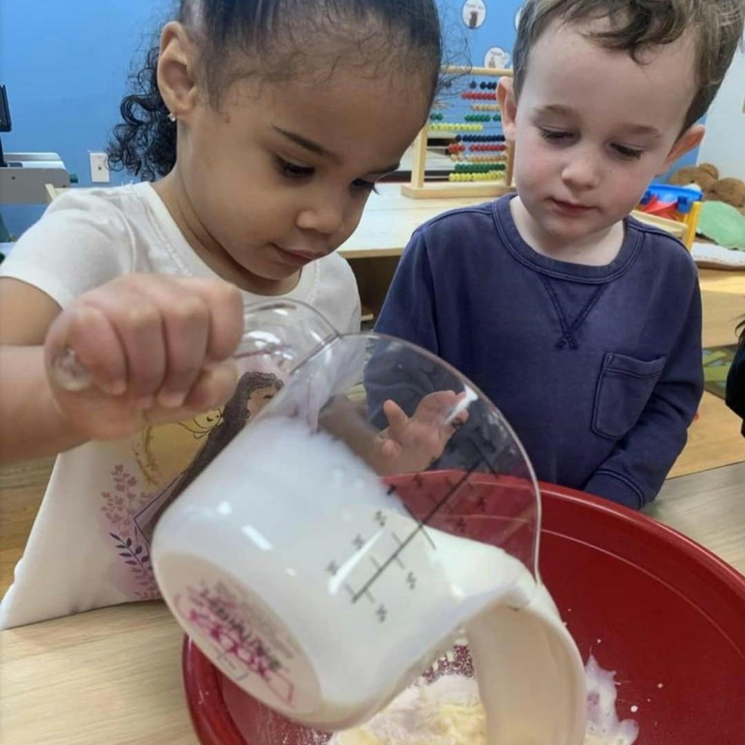 Two children pouring liquid from a measuring cup into a red bowl