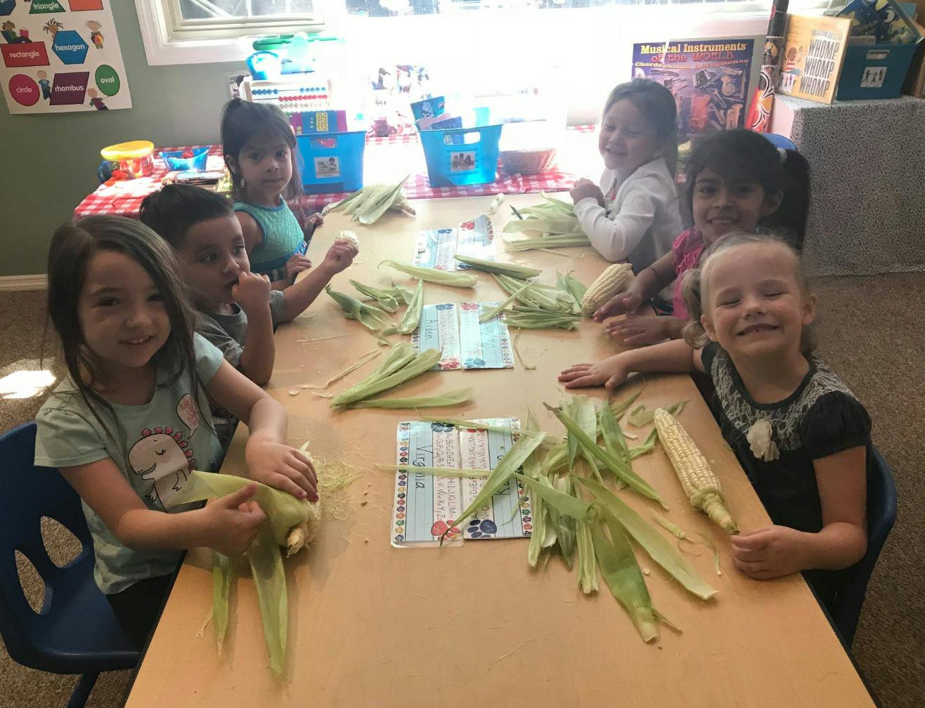 Children sitting at a table shucking corn