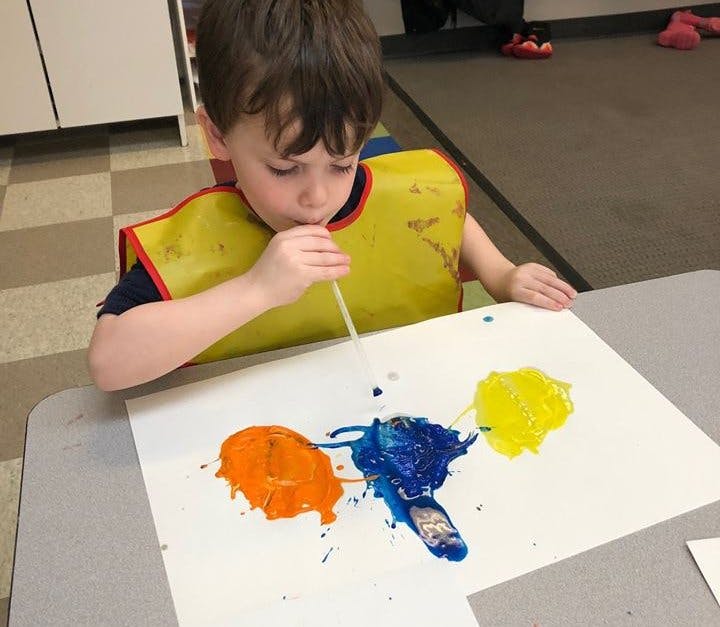 Child in a yellow smock blowing paint with a straw on paper