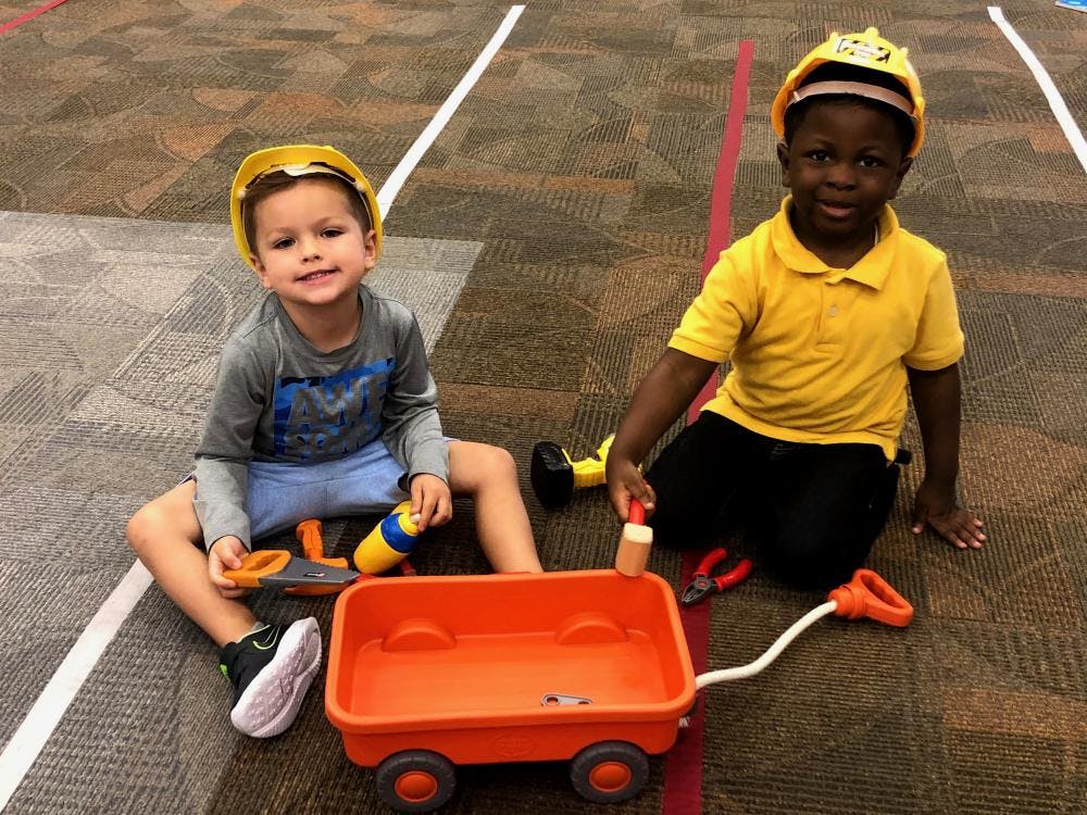Two children wearing hard hats playing with a toy wagon and tools