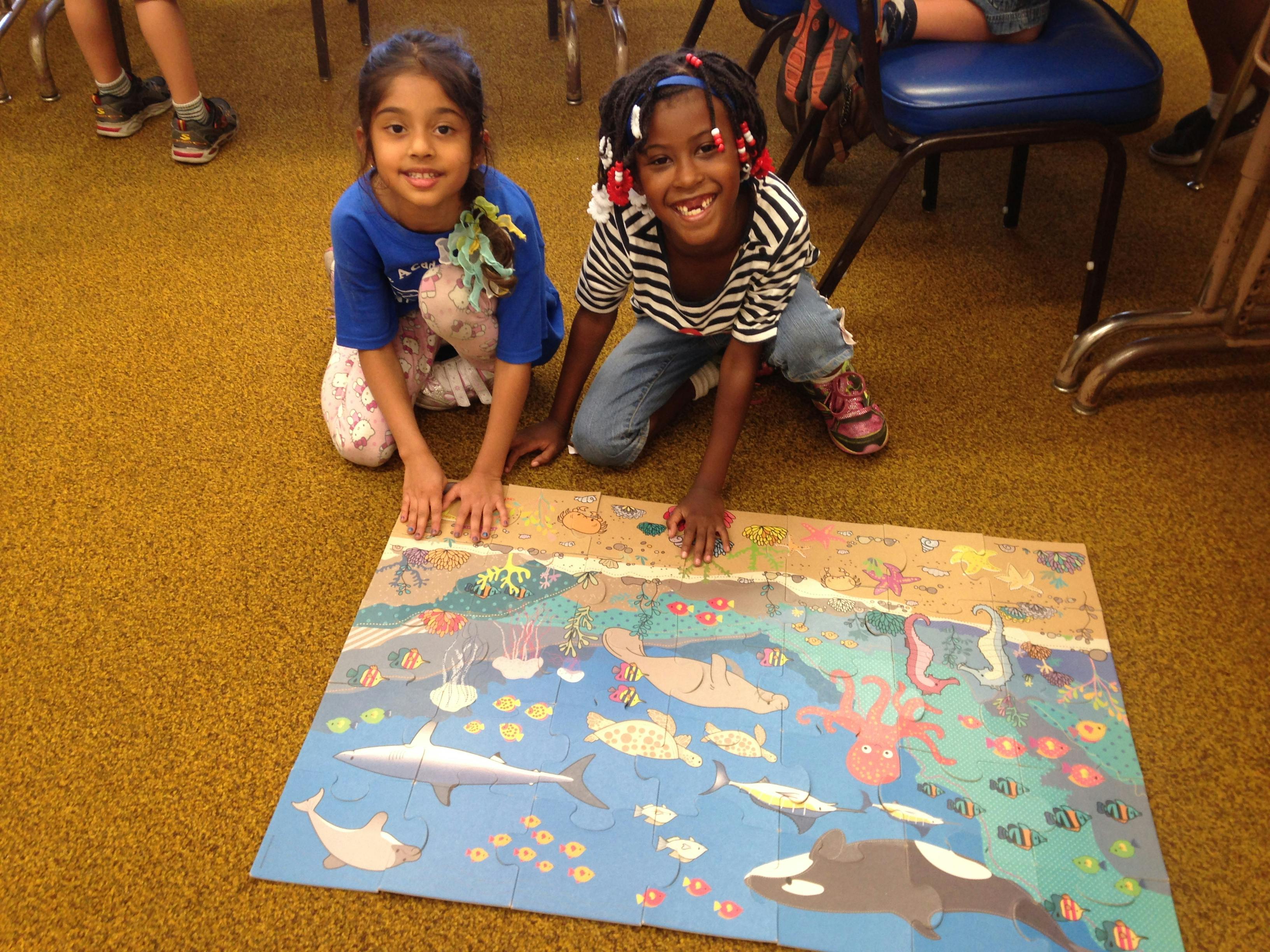 Two children kneeling on a carpet next to a large ocean-themed puzzle