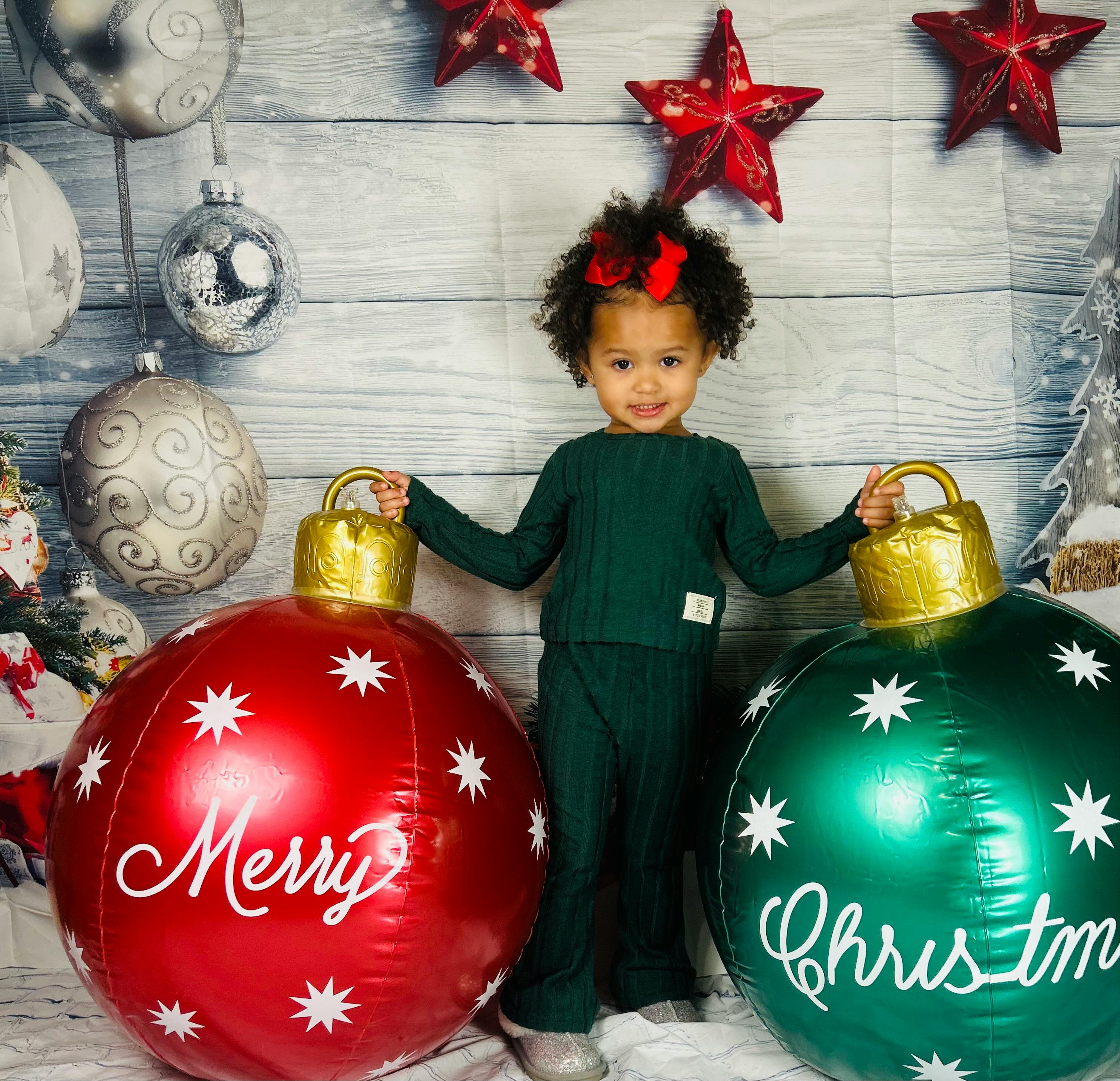 Child standing between two large Christmas ornaments, one says Merry, the other Christm