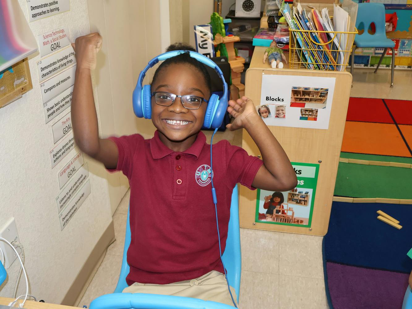 Child wearing headphones and glasses, raising arms in a classroom