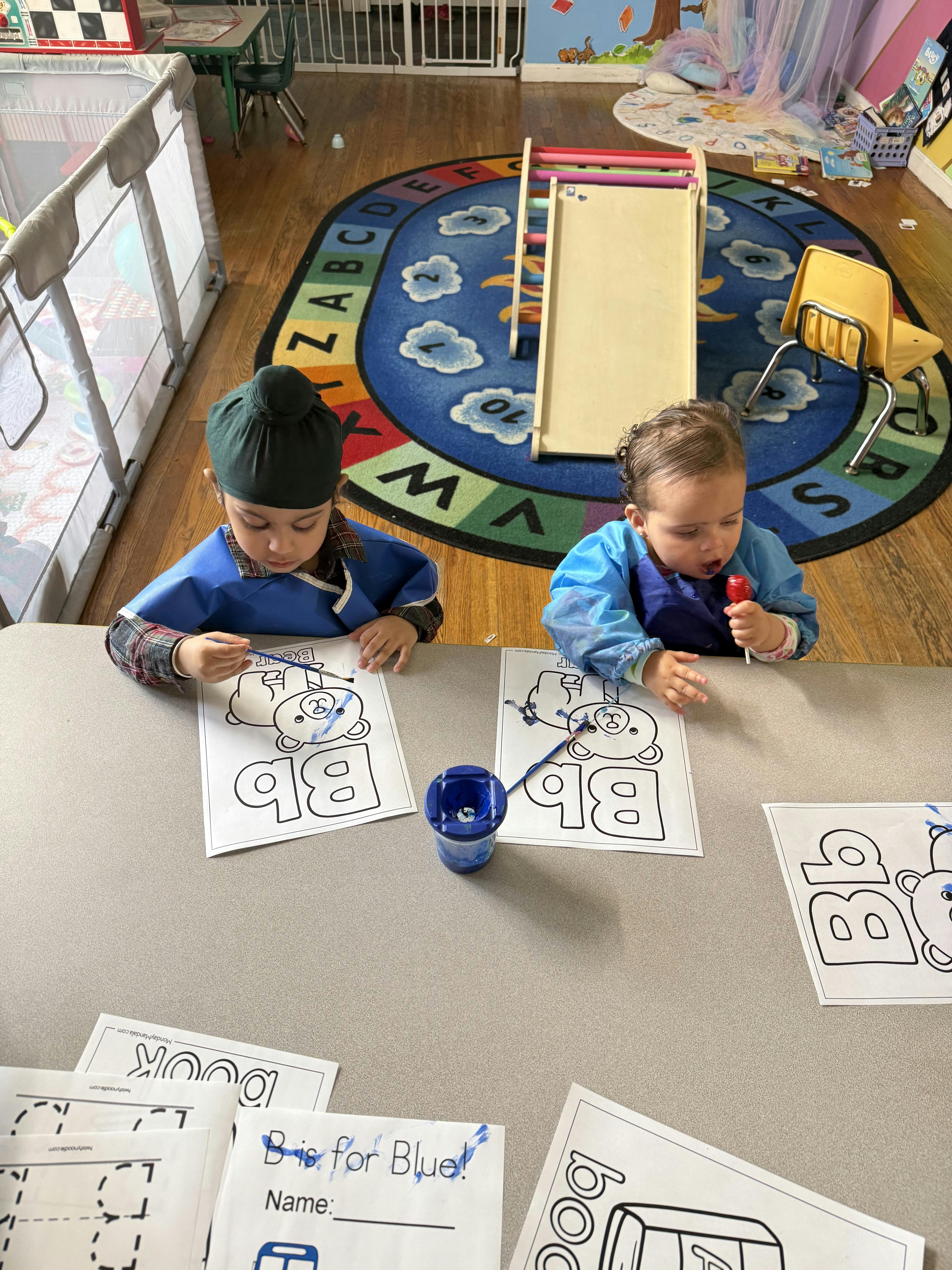 Two children painting bear coloring sheets at a table in a playroom