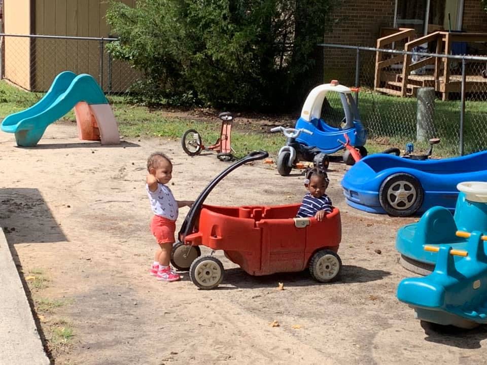 Child standing next to a wagon with another child sitting in it on a playground