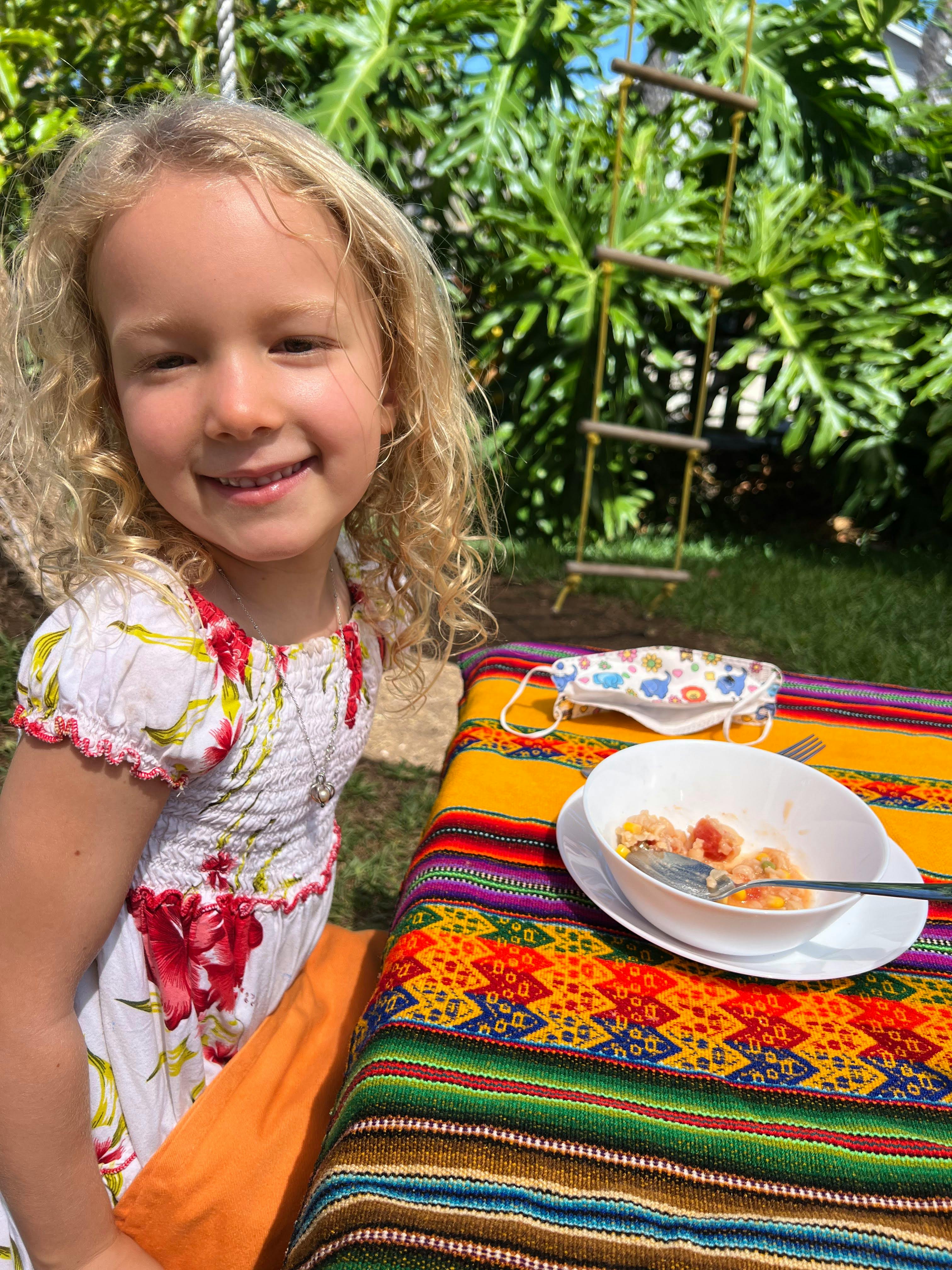 Child smiling outdoors next to a table with a bowl of food