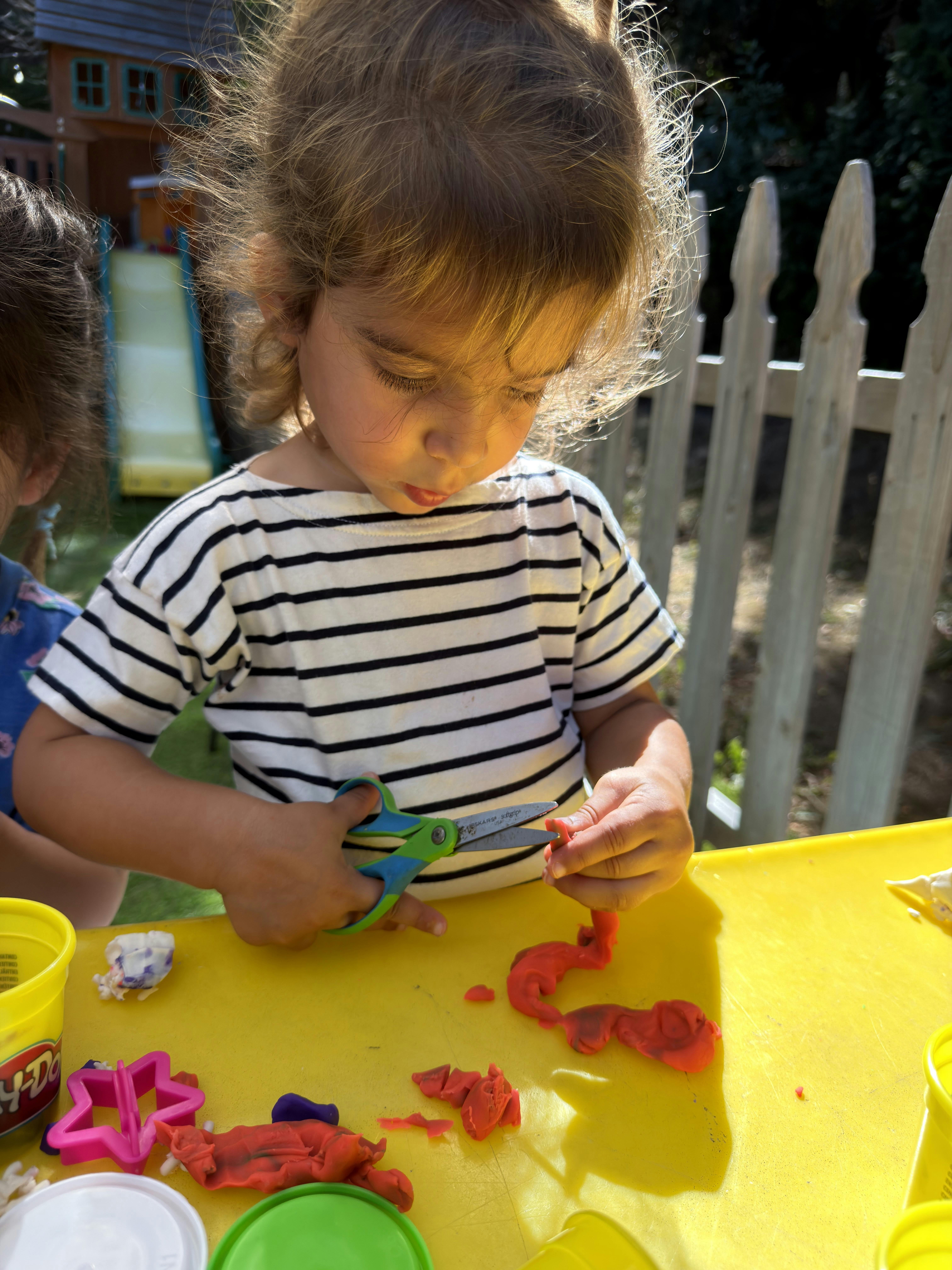 Child using scissors to cut red modeling clay at a table outdoors