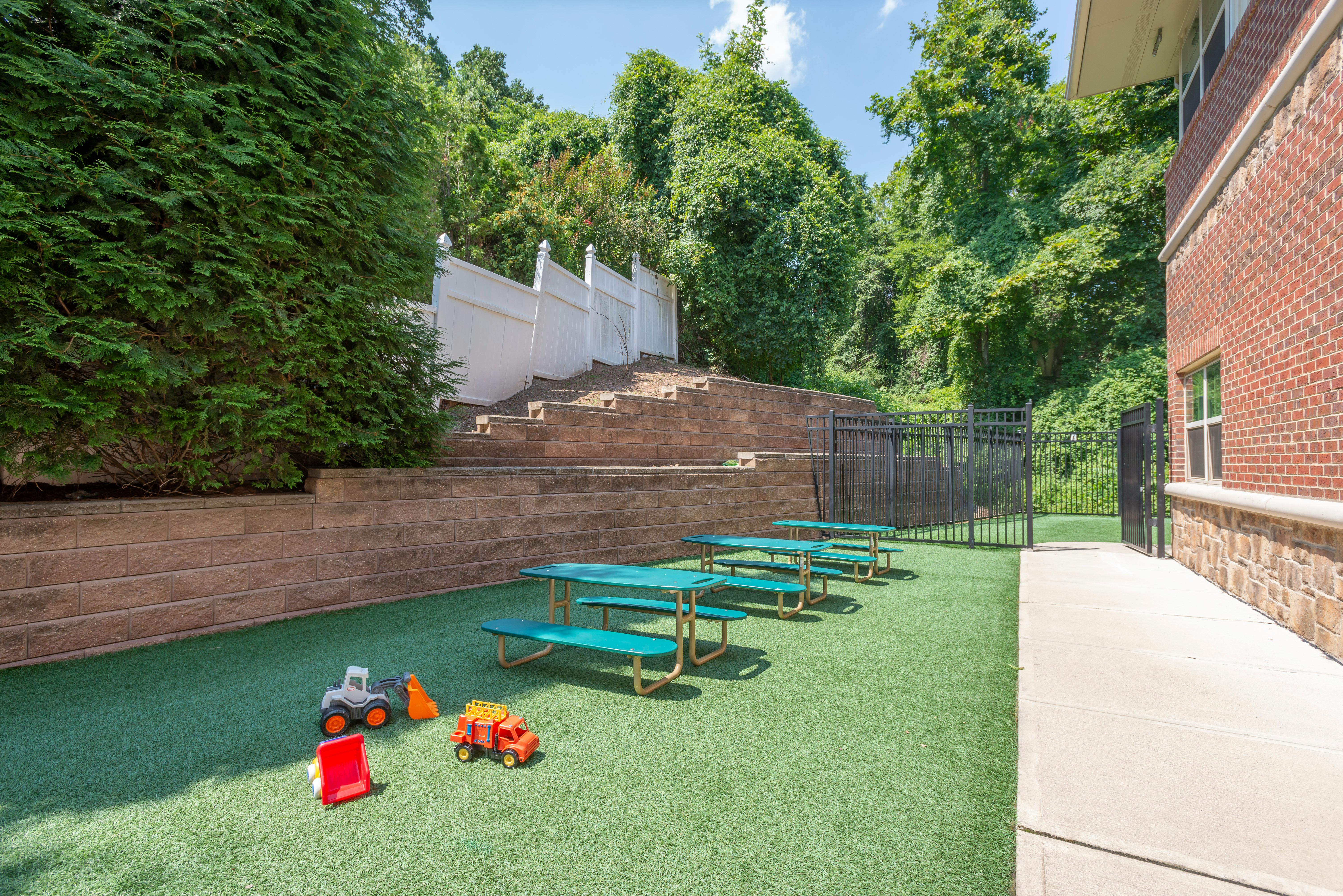 Outdoor play area with green turf, child-sized picnic tables, and toy trucks