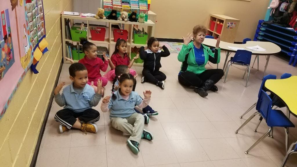 Adult and children sitting on floor with hands raised in a classroom