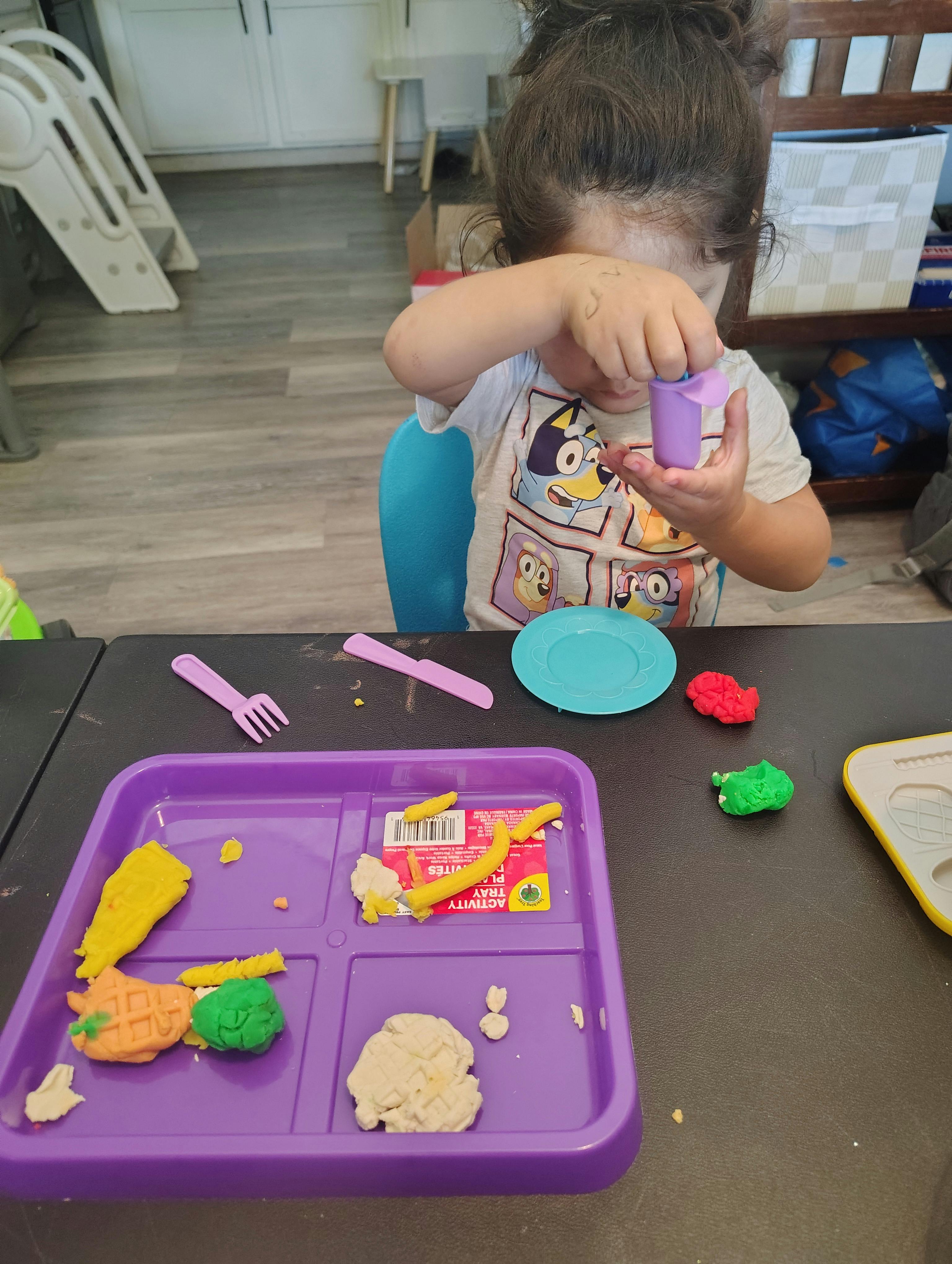 Child playing with modeling clay at a table with a tray and toy utensils