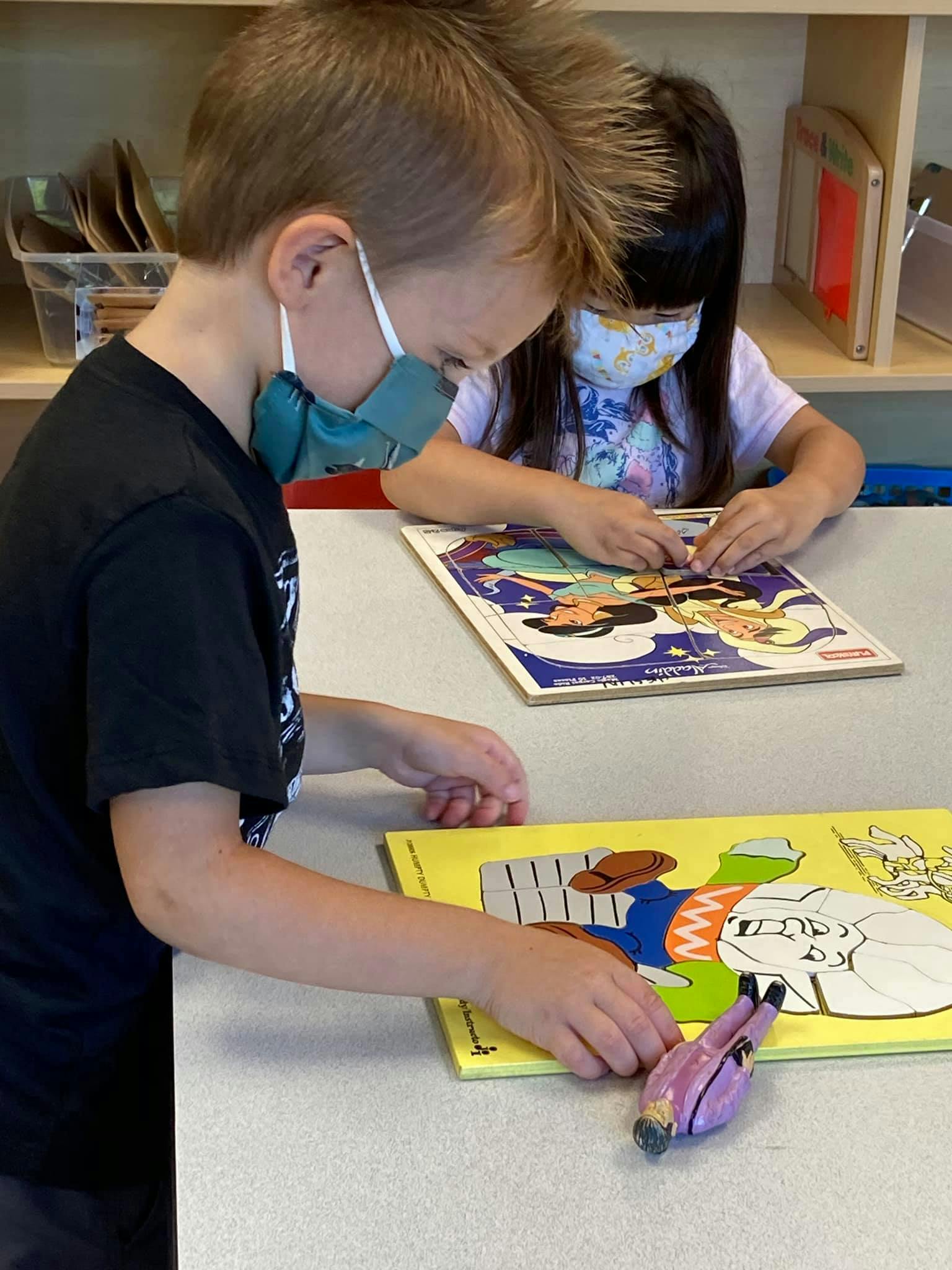 Two children wearing masks playing with puzzles at a table