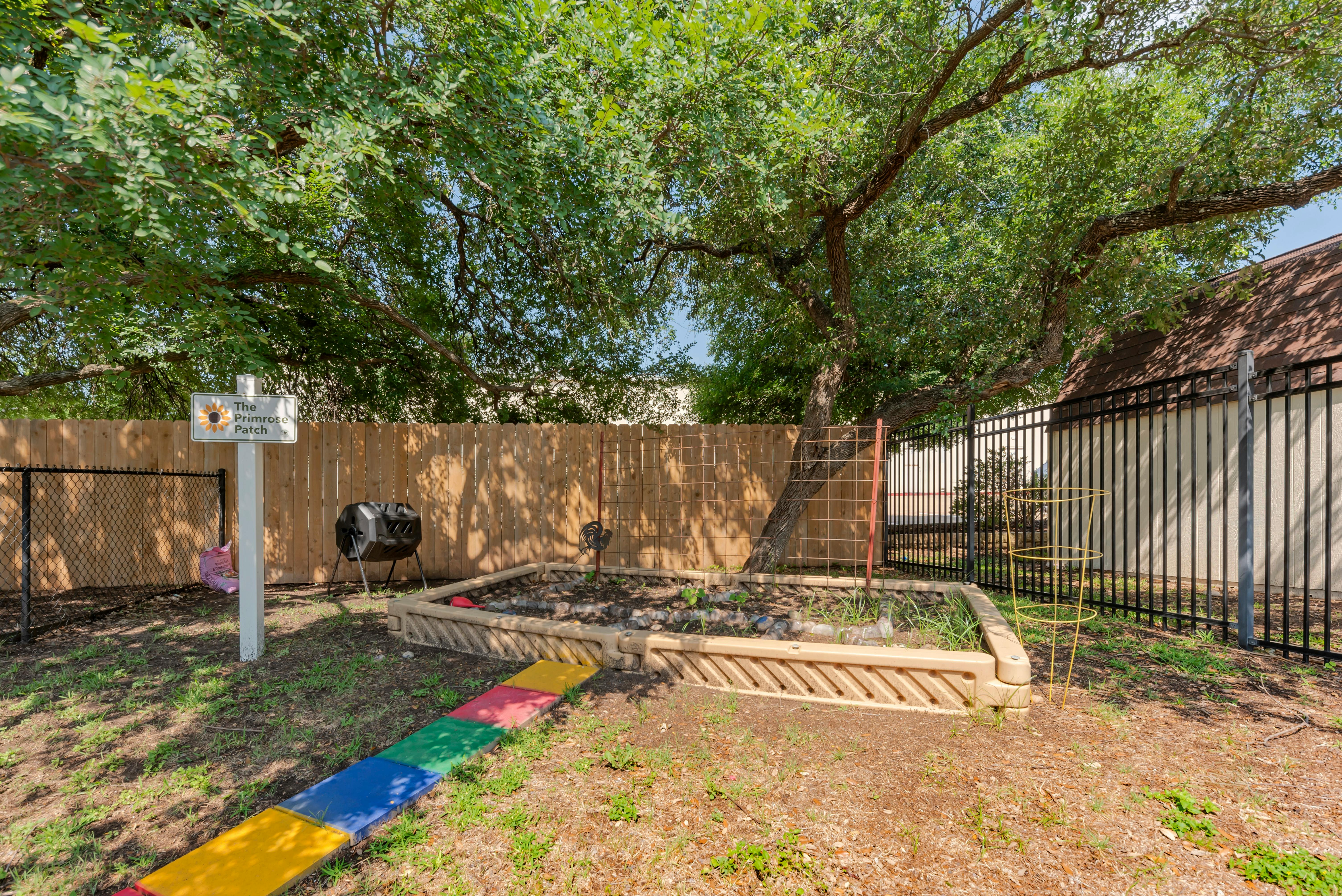 Raised garden bed and compost tumbler in a fenced outdoor area with large tree