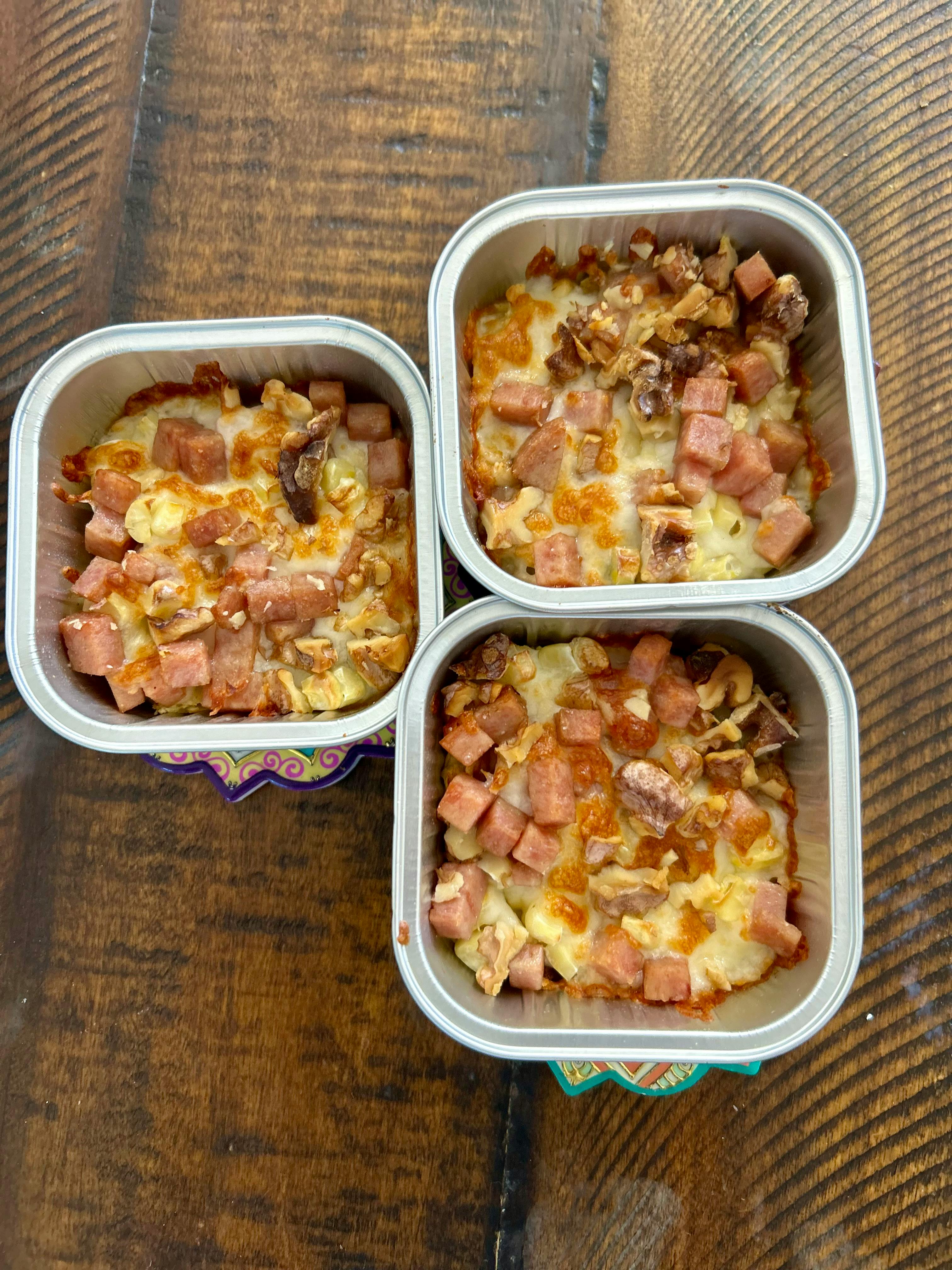 Three aluminum containers filled with baked food topped with ham and nuts