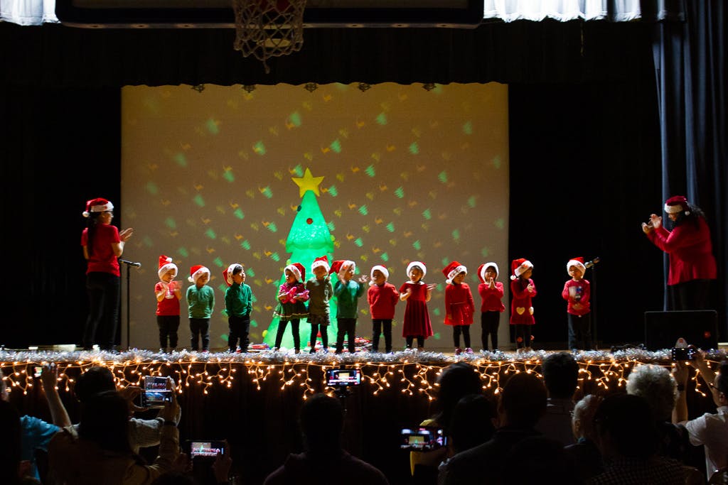 Children in Santa hats performing on a stage with a projected Christmas tree