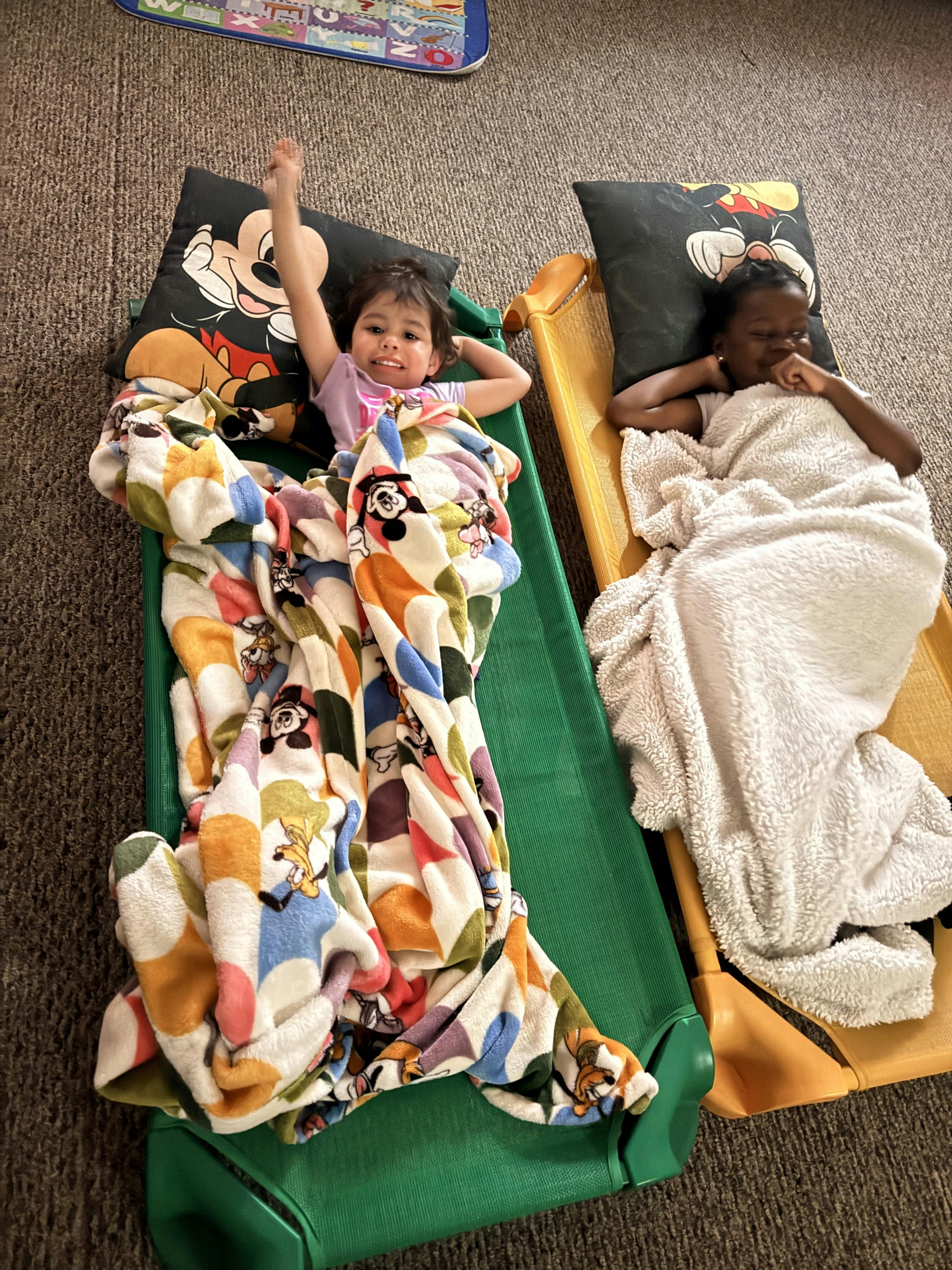 Two children lying on cots with blankets and pillows