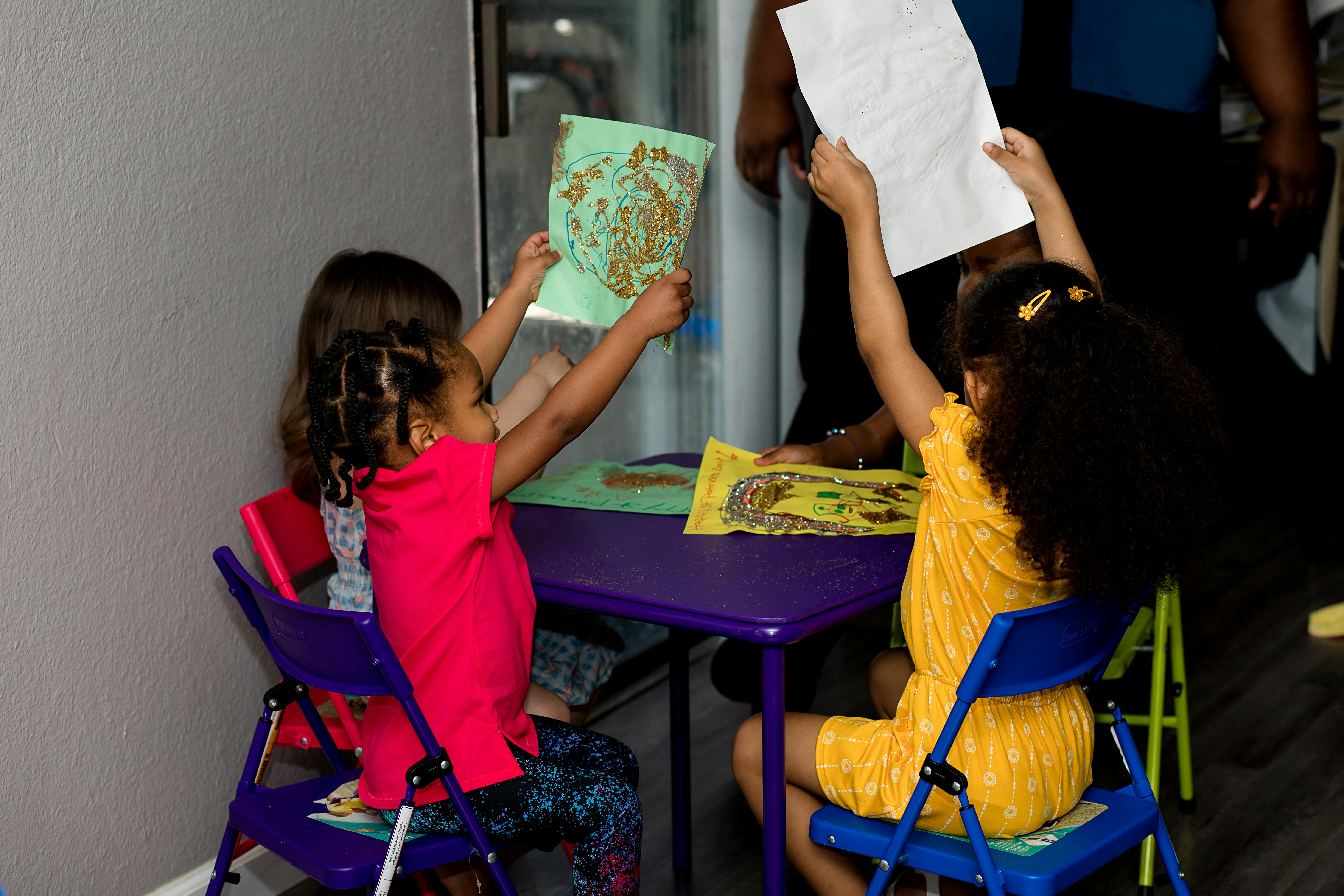 Two children sitting at a table holding up drawings