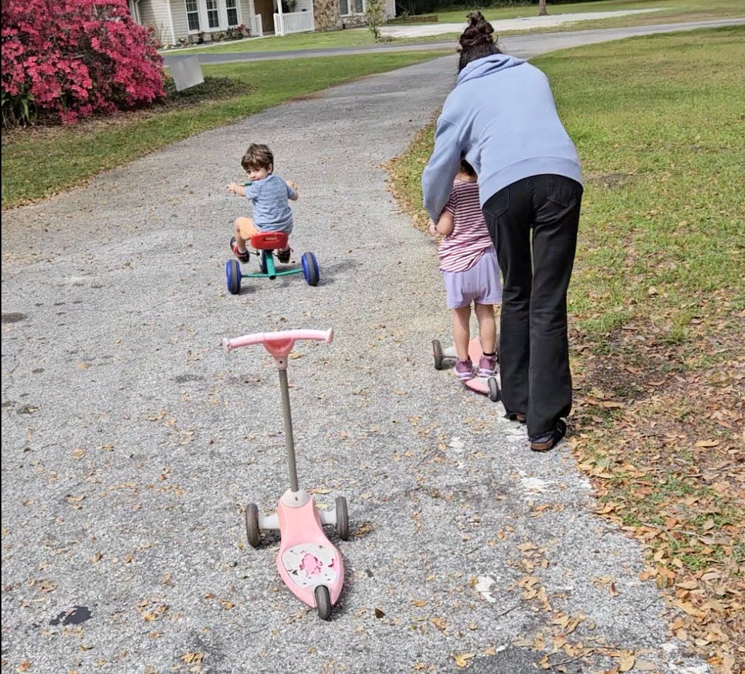 Person helping a child on a scooter with another child on a tricycle