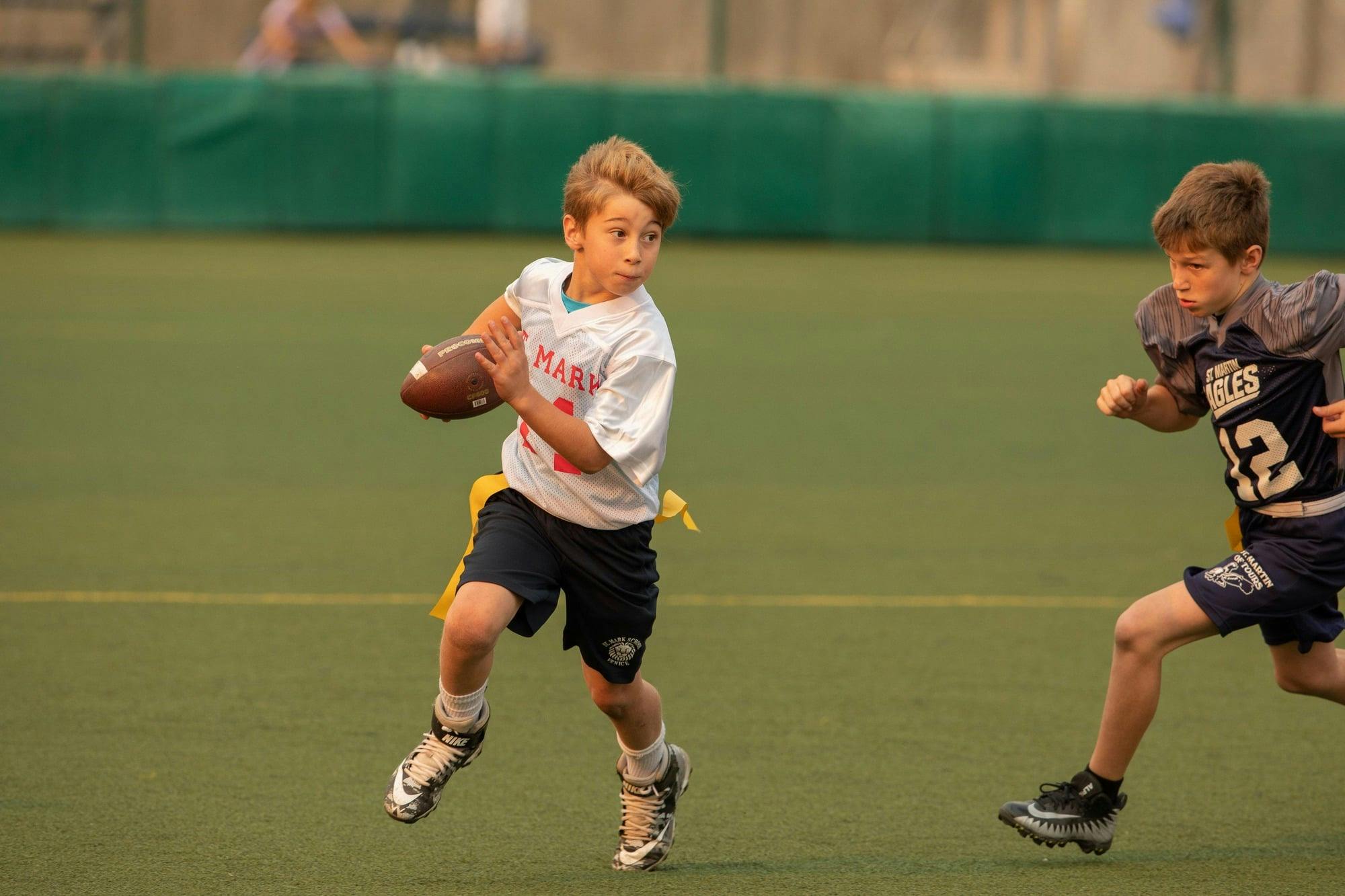 Child running with a football on a field, another child running behind them