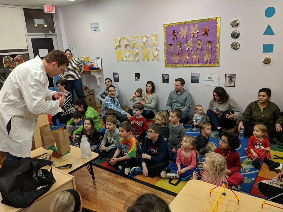 Person in lab coat demonstrating to children and adults in a classroom