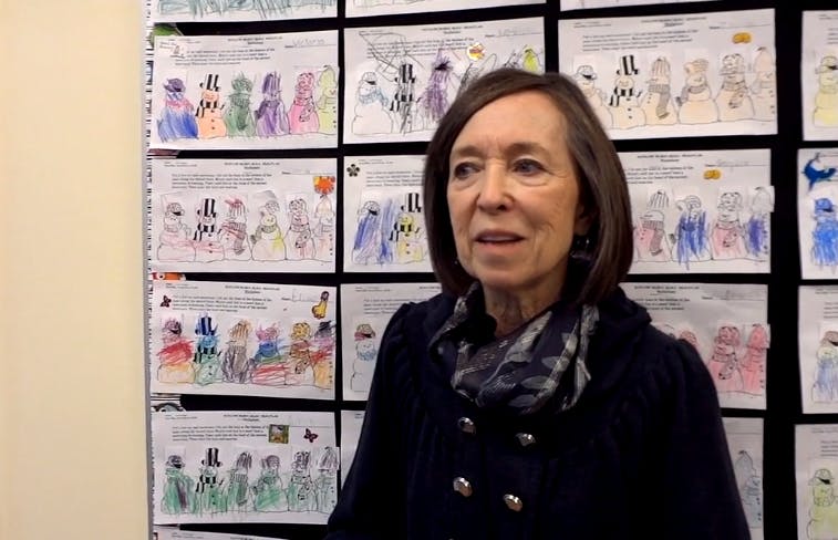 Person in a dark coat and scarf standing in front of children's drawings