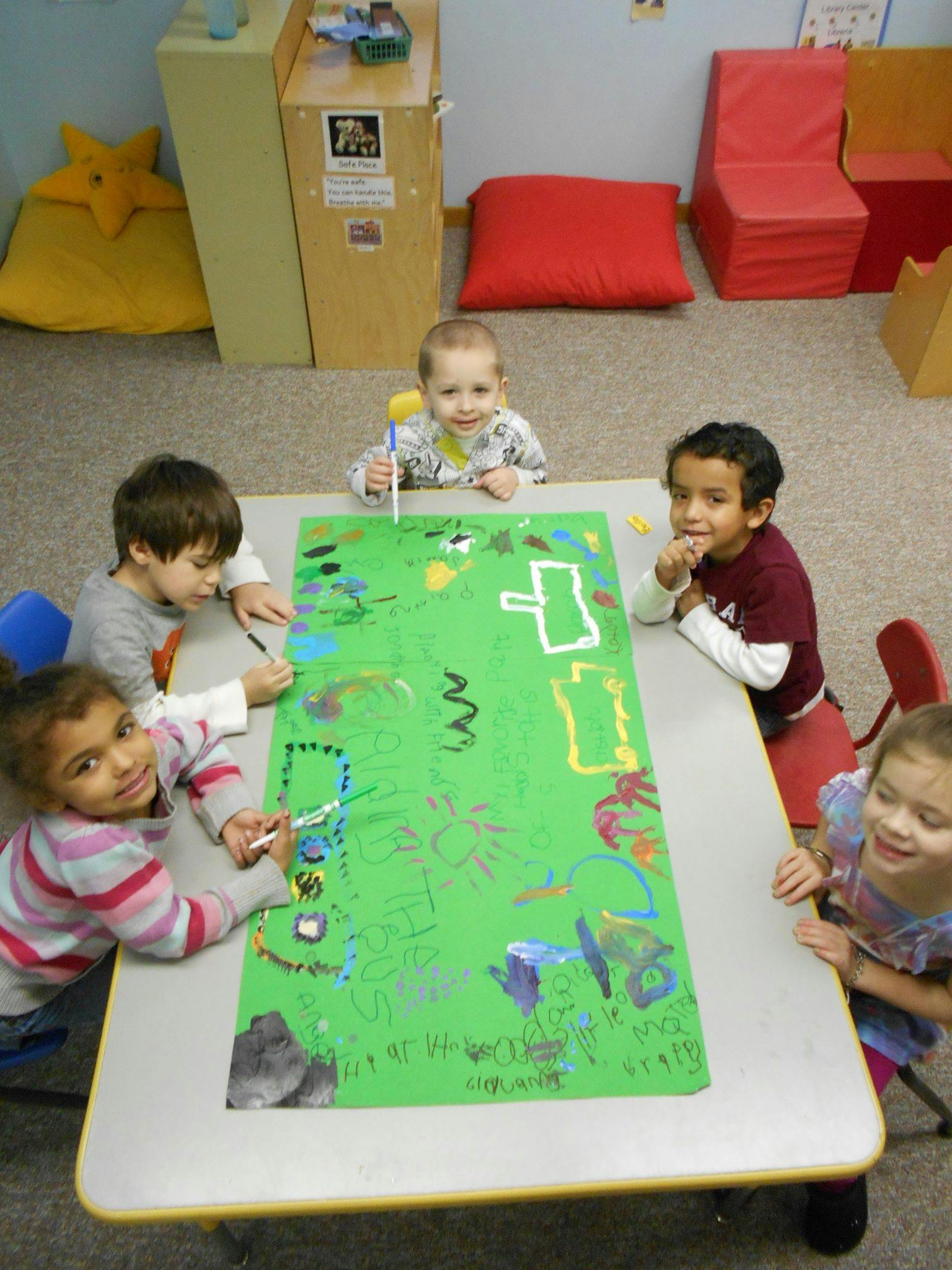 Four children drawing on a large green paper at a table in a classroom
