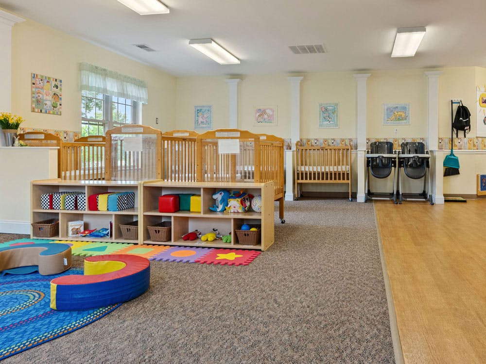 A brightly lit nursery room with several wooden cribs and shelves of toys