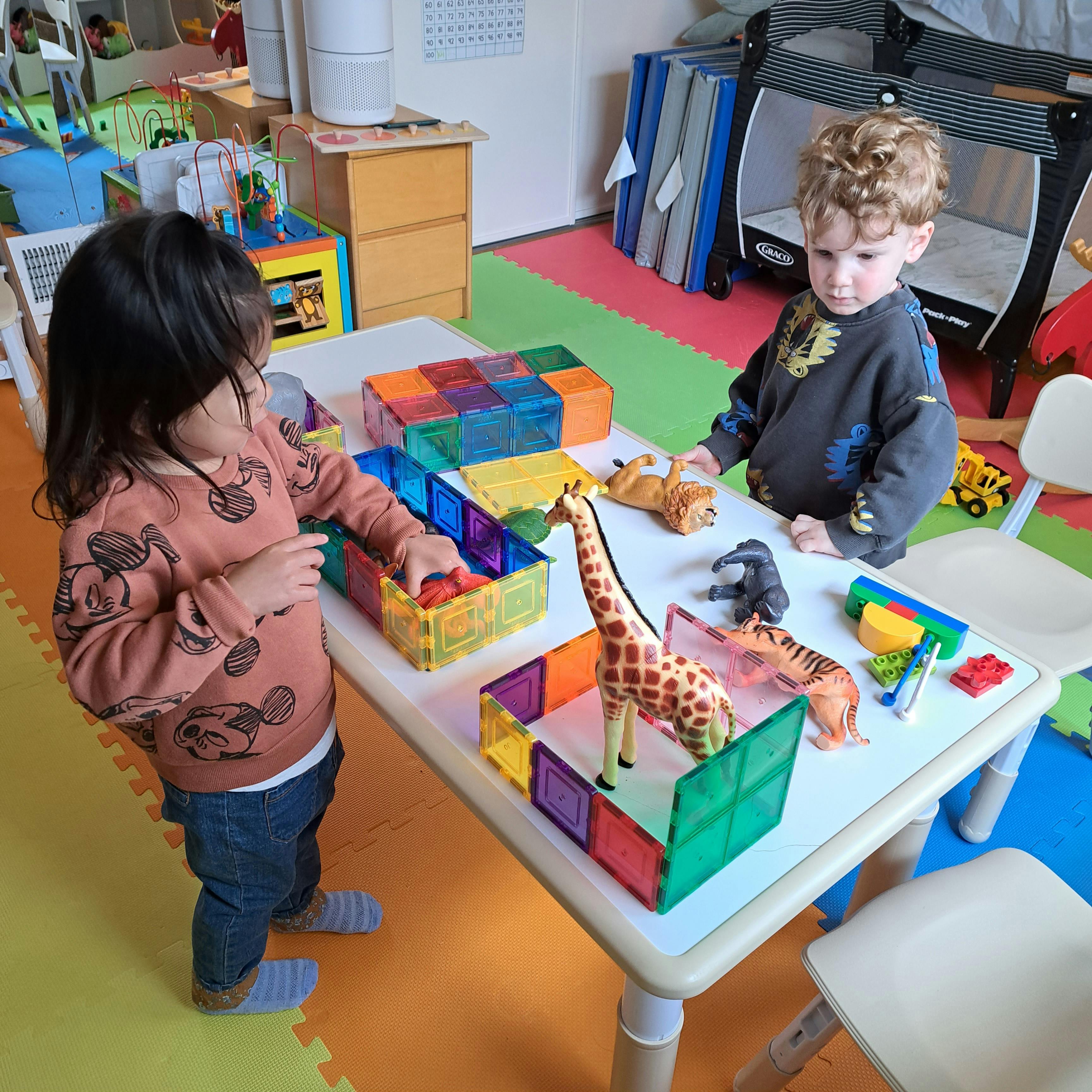 Two children playing with magnetic tiles and animal figures at a table