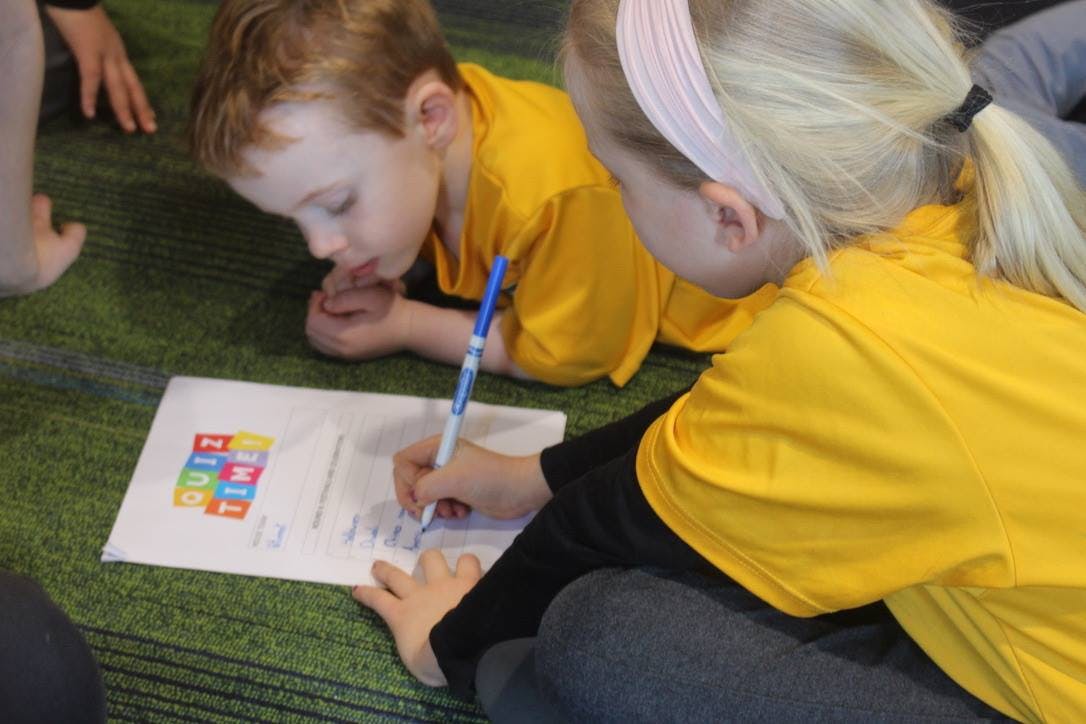 Two children in yellow shirts writing on paper on the floor