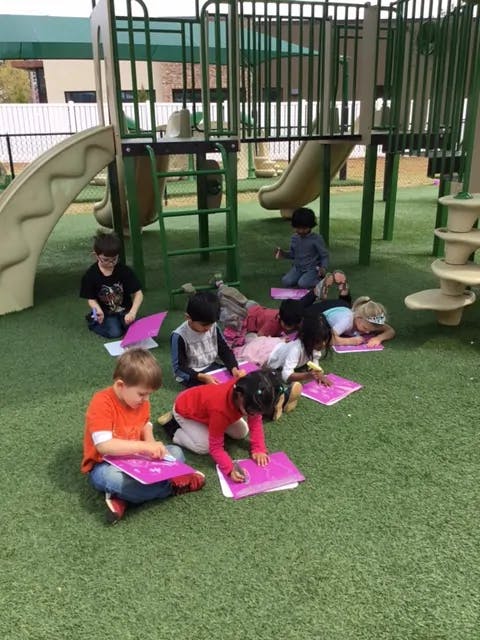 Children sitting on artificial grass writing on pink paper near playground equipment
