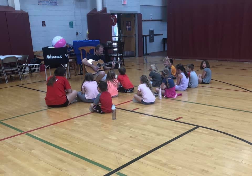 Person playing guitar for children sitting on a gymnasium floor