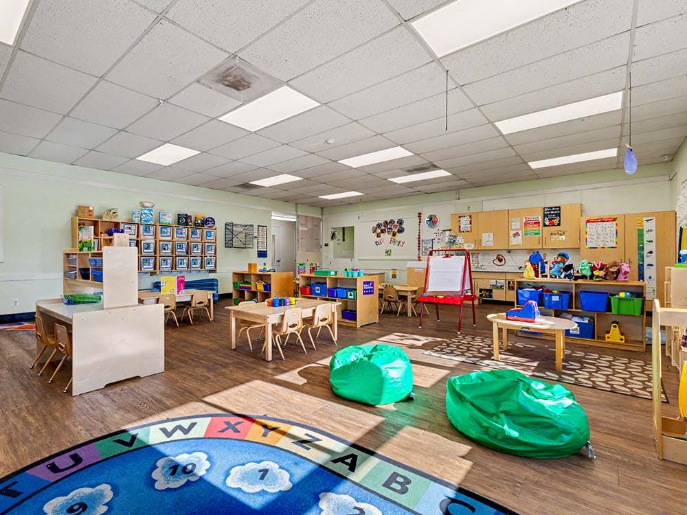 Empty classroom with small tables and chairs, shelves with toys, and an alphabet rug