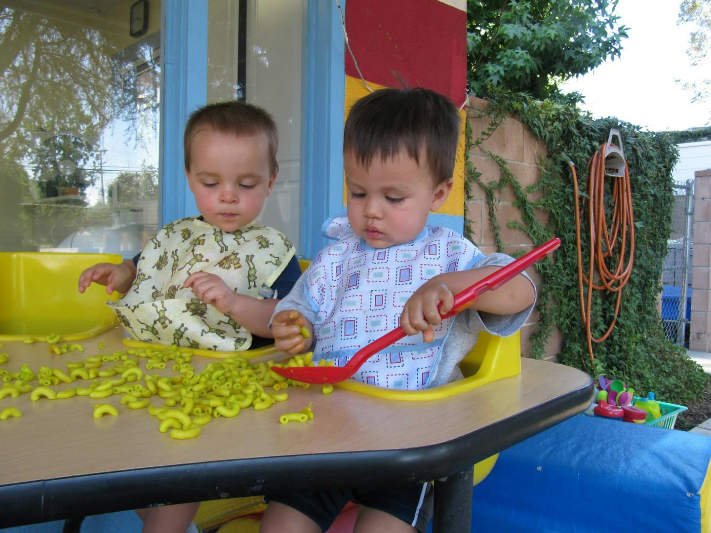 Two children playing with yellow pasta shapes and a red spoon at a table