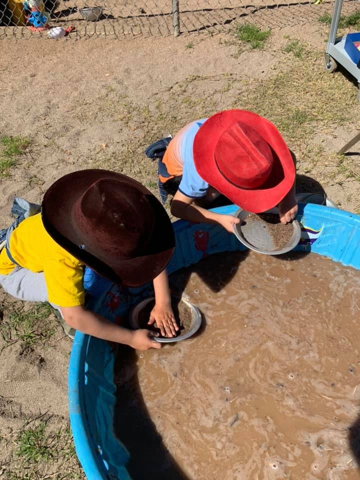 Two children in hats playing in a mud puddle with pans