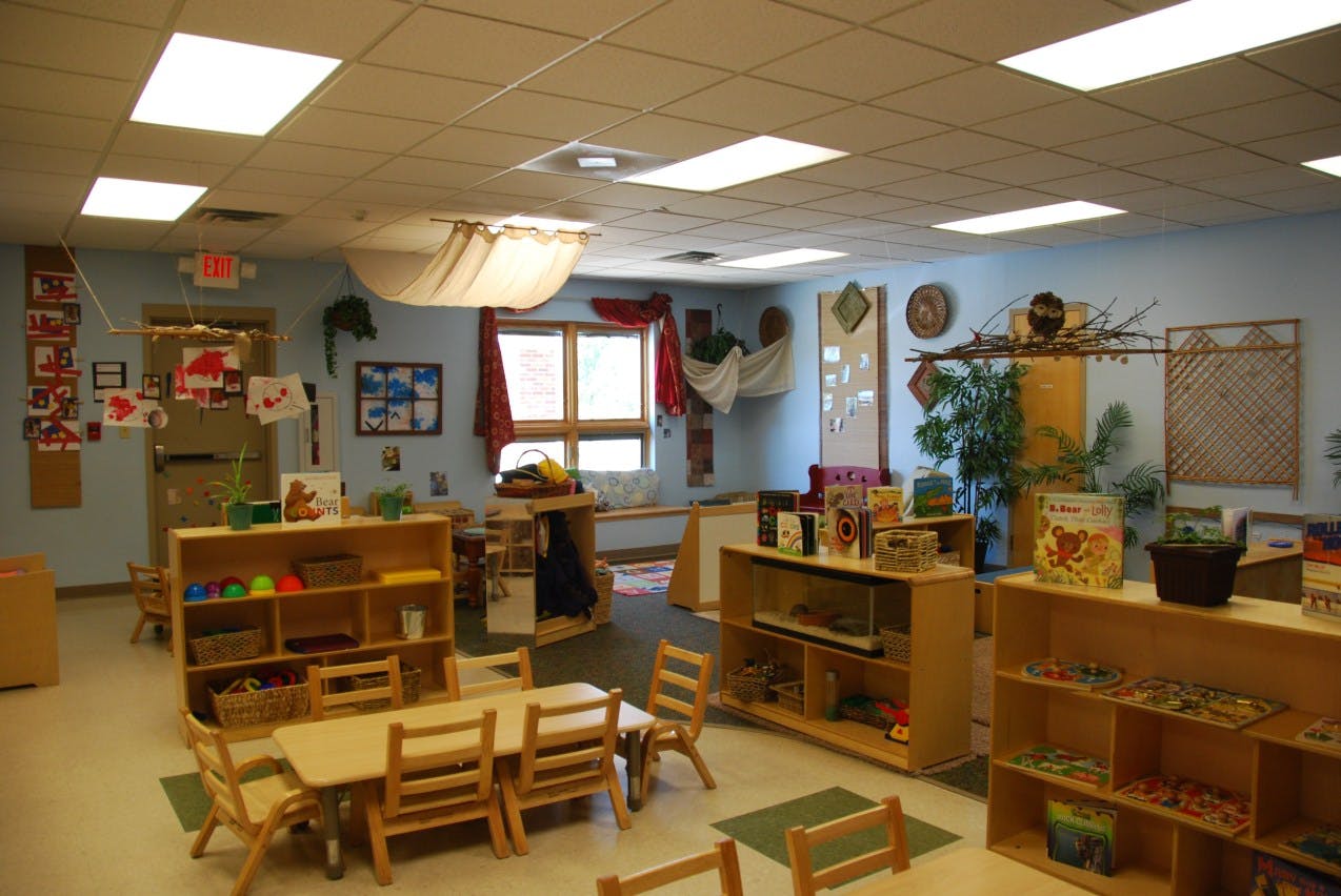 Classroom with small wooden tables and chairs, shelves with learning materials