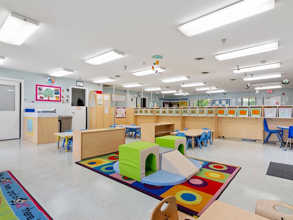 A classroom with small tables, chairs, and play equipment on a colorful rug