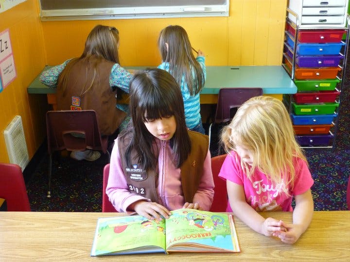 Two children reading a book at a table, with other children in the background