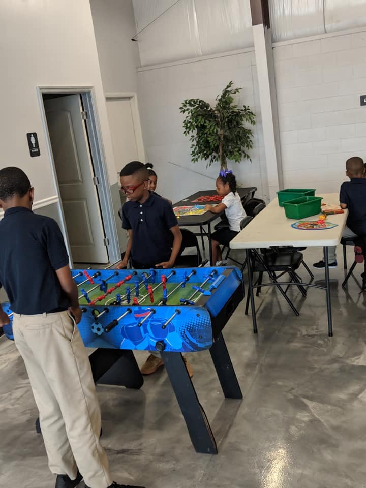Two children playing foosball in a room, other children at tables