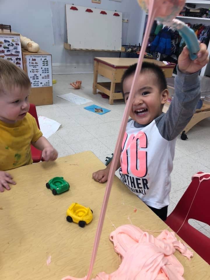 Child holding up pink slime in a classroom with another child at a table
