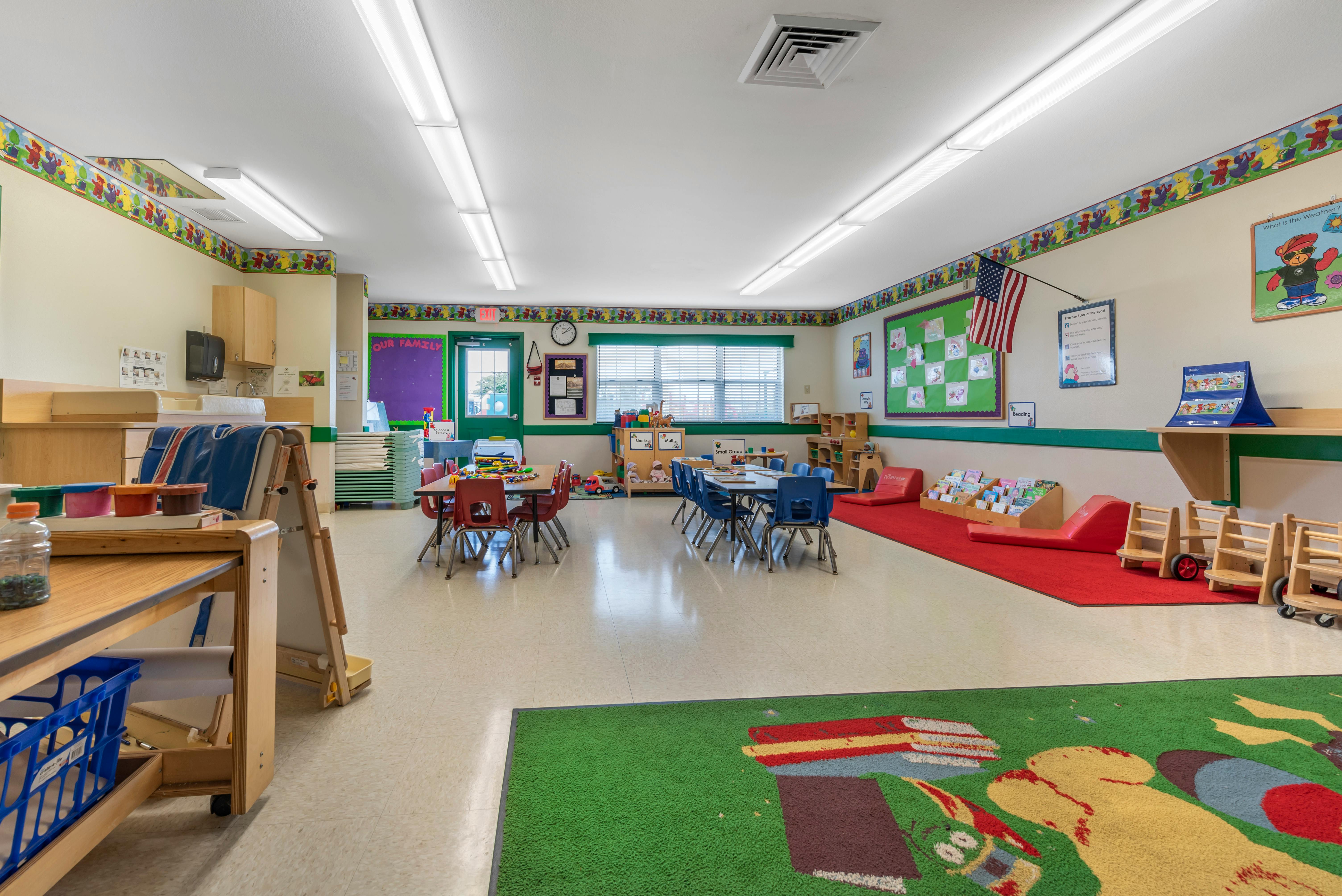 Classroom with small tables and chairs, a play rug, and shelves