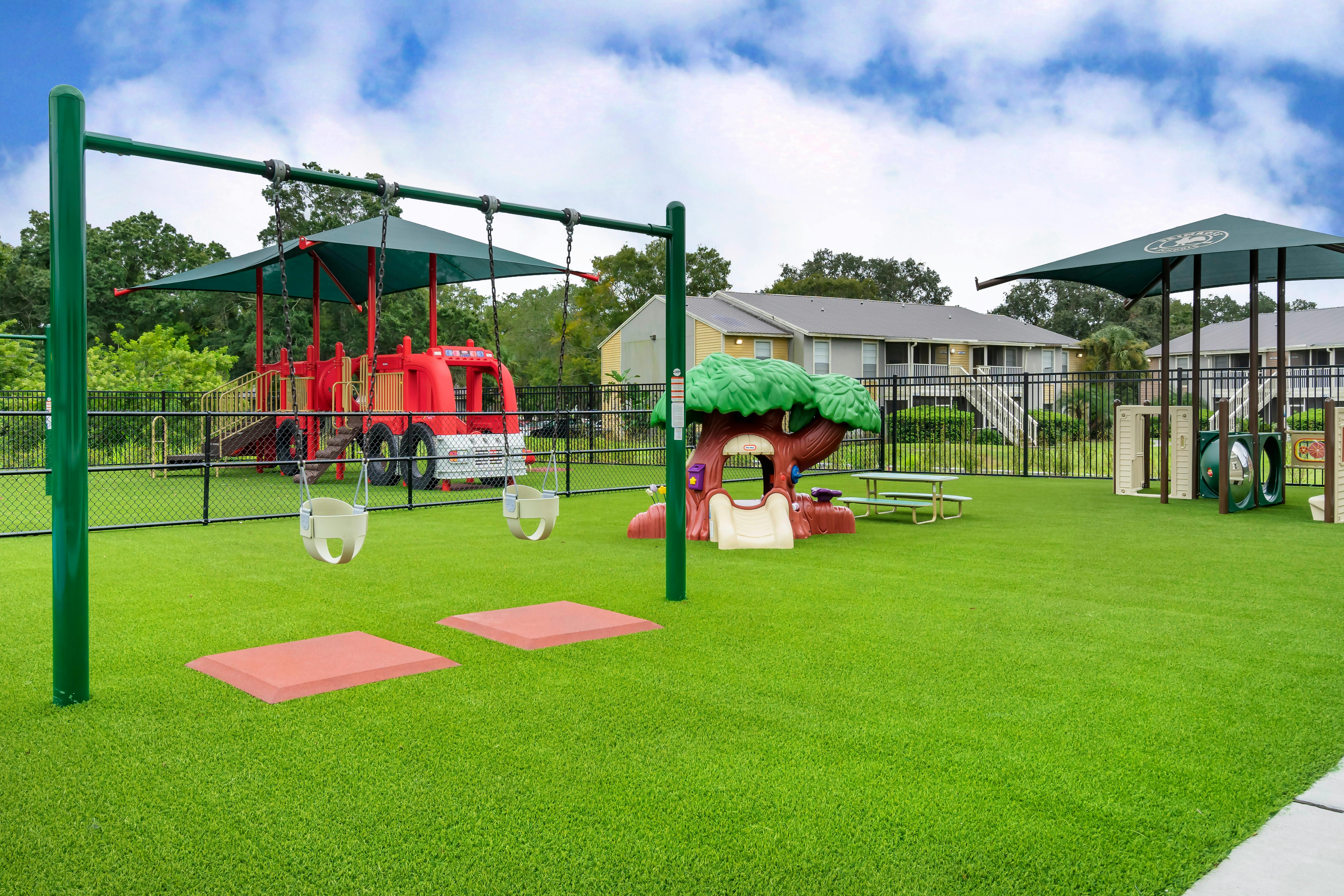 A fenced playground with green turf, swings, and play structures