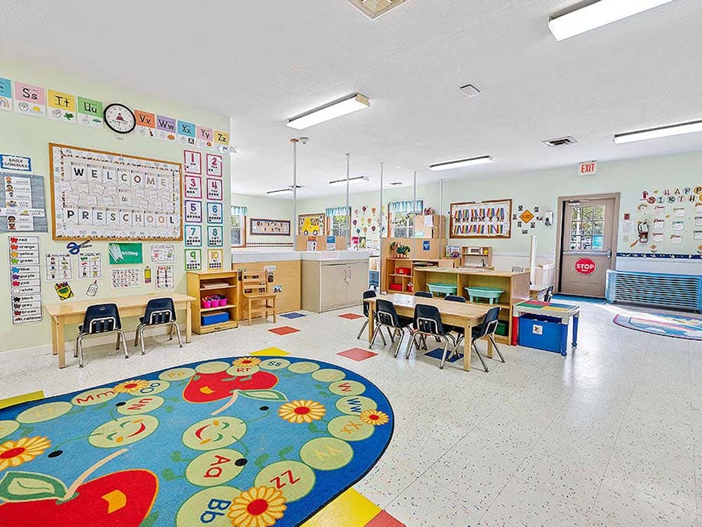 Empty preschool classroom with small tables, chairs, and a colorful rug