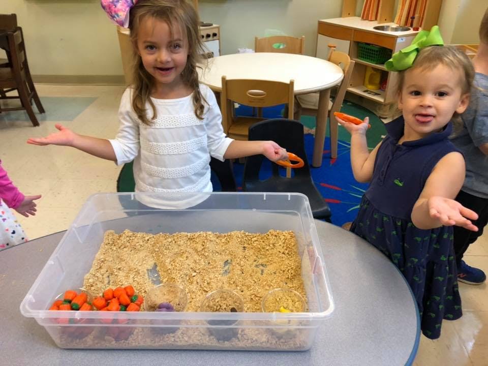 Two children standing by a sensory bin with rice and small objects