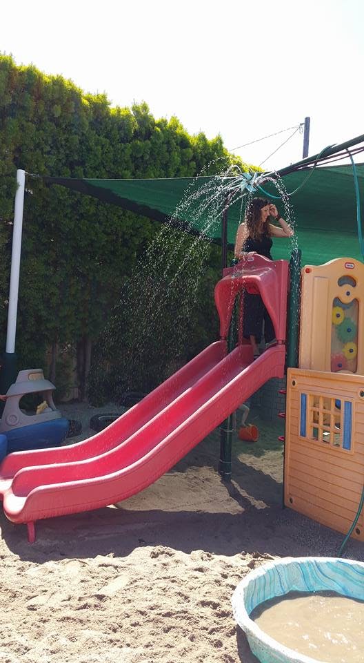 Person standing on a red playground slide with water spraying overhead