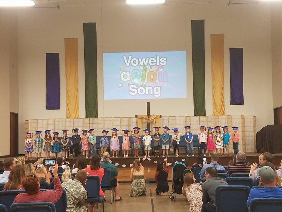 Children in graduation caps on a stage with a screen displaying "Vowels Song"