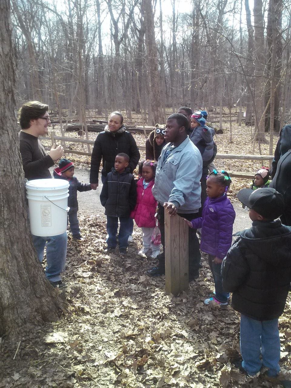 Person speaking to a group of adults and children in a wooded area