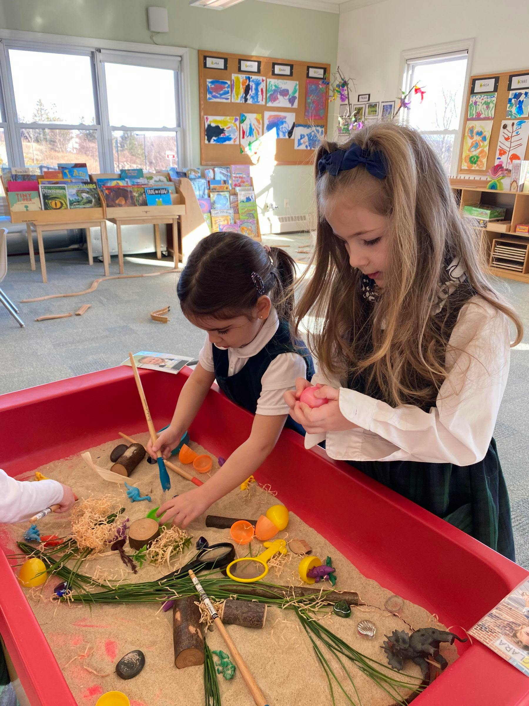 Two children playing with sand and natural elements in a red sensory bin
